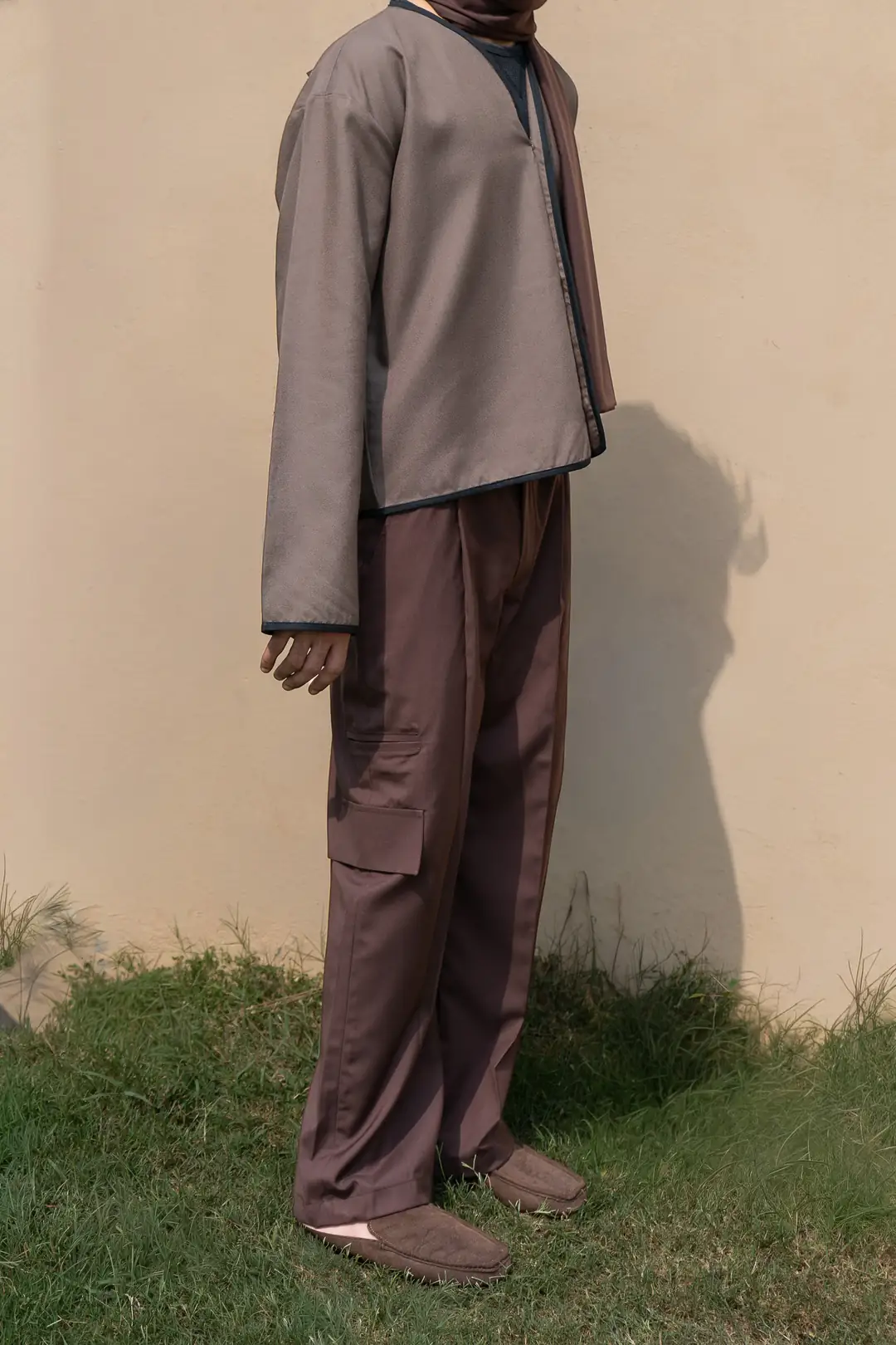 A man in a taupe collarless overshirt with black piping, brown pleated cargo trousers and brown suede loafers